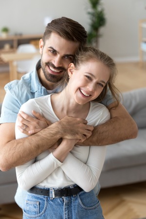 Portrait of happy husband hold in arms young wife posing at home, excited millennial couple hugging looking at camera making family picture in living room, loving man and woman embrace laughingの写真素材
