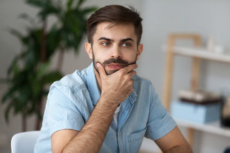 Close up of pensive millennial man sit at office home desk thinking of problem solution, thoughtful male lost in thoughts ponder or consider plan implementation, guy look in distance making decisionの写真素材