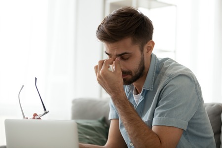 Tired millennial man massage nose bridge having health problems, suffer from headache, exhausted male use laptop take glasses off having blurry vision or dizziness working too long at computerの写真素材