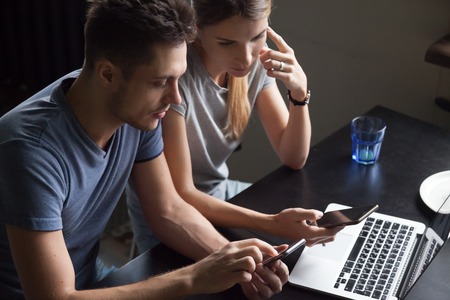 Serious young couple using smartphones and laptop, reading bad news, email, using mobile apps, searching information online, holding phones in hands, checking bank accountの写真素材