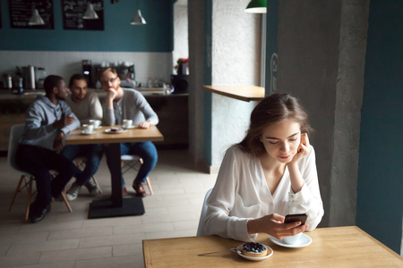 Diverse young guys interested with beautiful girl sitting at near table using smartphone, enjoying coffee in cafe, curious males looking at pretty female, liking or falling in love from first sightの写真素材