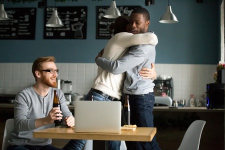 Happy multiethnic friends hug celebrating team victory hanging out in pub, excited diverse males embrace congratulating with goal score, guys have fun drinking beer watching football on laptop in cafeの写真素材