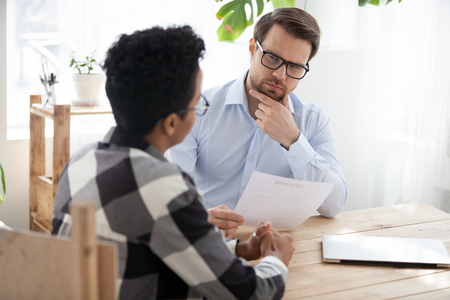 Suspicious male employer look at African American millennial applicant, doubt in candidature, thoughtful HR manager consider resume hesitating about black candidate experience at office interviewの写真素材