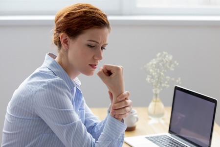 Businesswoman sitting at desk opposite pc suffers from pain in hand while working in office. Young female massaging wrist having carpal tunnel syndrome, caused by long-term use of keyboard and mouseの写真素材