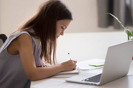Young girl, intern holding pen in hand, making notes, working with laptop in office, female student preparing for test or exam, writing essay in notebook, studying in college, universityの写真素材