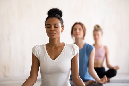 Group diverse young beautiful women sitting in lotus position meditating during session at yoga studio. Girls practising exercises visualizing calming the brain increasing awareness and attentivenessの写真素材