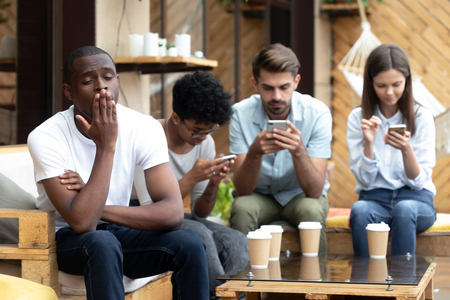Bored African American man sitting with people with phone dependence, yawning, friends using mobile devices, apps, looking at screen, ignore each other in cafe, smartphone addiction conceptの写真素材