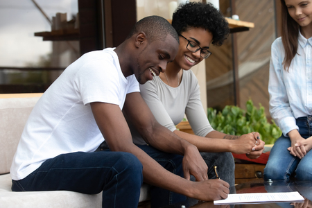 Happy African American couple signing contract, making agreement with businesswoman, female realtor, wedding planner, financial advisor, smiling clients with seller in cafe, successful negotiationsの写真素材