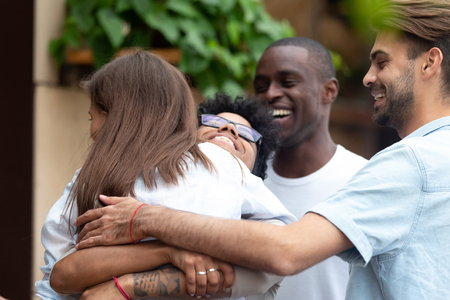 Happy meeting of two female best friends after long time embracing in cafe, two young multiracial women hugging each other, excited men supporting, good multiethnic friendship conceptの写真素材