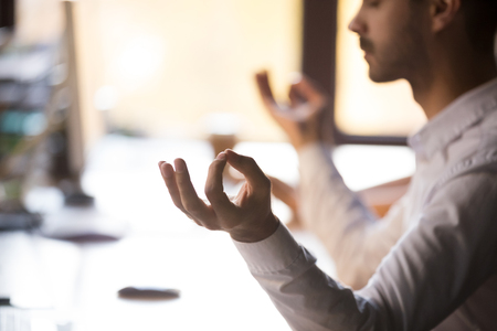 Close up view of mindful calm man sitting at home office desk holding hands in mudra doing yoga exercise for relaxation concentration at workplace, meditation at work, no stress free relief conceptの写真素材