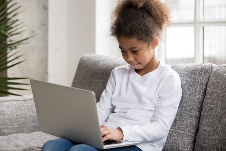 Curious preschool african mixed race girl using laptop on couch, little smart black kid typing on computer chatting with friends online alone at home, child security, children and gadget conceptの写真素材