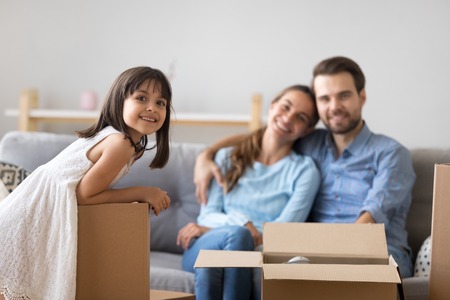 Portrait of happy kid girl posing with cardboard boxes on moving day with smiling parents at background, cute little child daughter looking at camera in new home enjoying family relocation conceptの写真素材