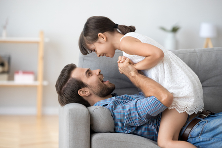Happy dad and playful cute kid girl holding hands playing on sofa at home, father and little smiling daughter having fun relaxing spending time together, daddy and funny child enjoy communicationの写真素材