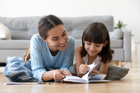 Caring smiling mother reading book with little kid girl lying on warm floor at home, mom or baby sitter playing having fun telling fairy tale to child daughter, underfloor heating, family activitiesの写真素材