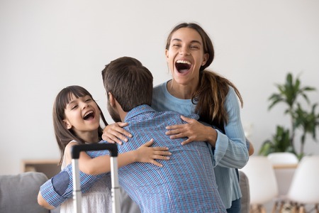 Excited wife and kid daughter hugging father arriving returning after long trip with suitcase, happy dad embracing surprised loving family welcoming missing daddy coming back home, reunion conceptの写真素材