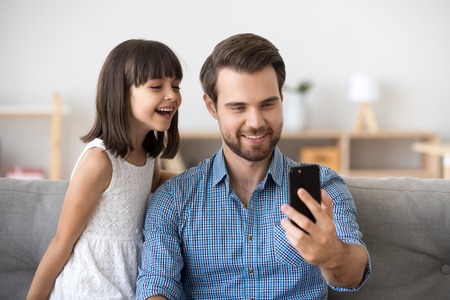 Happy dad laughing taking selfie with kid daughter on smartphone at home, smiling daddy and child making photo or call looking at cellphone screen, father and cute girl watching funny video on phoneの写真素材