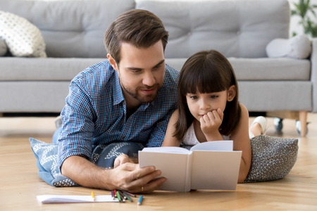 Cute little girl listening to dad reading fairy tale lying on warm floor together, caring father holding book teaching child daughter spending time with focused kid, family hobbies activities at homeの写真素材