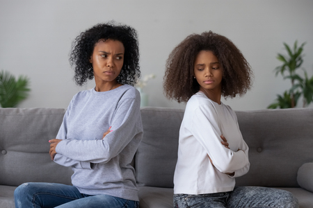 Angry African American mother and daughter sit back to back on couch, avoid talking or looking, mad black mom and teenage girl have fight or quarrel, stubborn parent and kid refuse to compromiseの写真素材