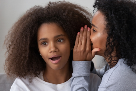 African American female tell secret to surprised girlfriend, black young mom or nanny spend time together with teenage girl, sharing or gossiping, modern parent and teen daughter best friends conceptの写真素材