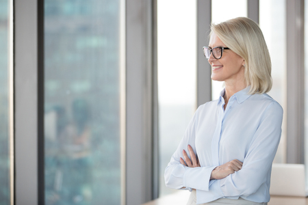 Smiling successful middle aged businesswoman looking through window dreaming of new goal opportunity in modern luxury office, serious mature senior old woman planning future, business vision conceptの写真素材