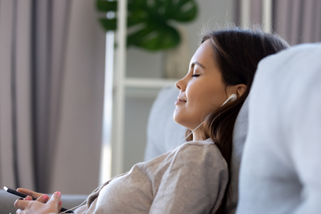 Serene calm young woman wearing earphones relaxing on sofa listening music on smartphone player app, happy teen girl in headphones having peaceful mood holding mobile phone chilling lounging at homeの写真素材