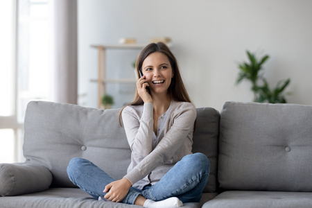 Happy young woman caller talking on the phone at home, cheerful teen girl enjoys pleasant mobile conversation, smiling millennial female holding cell speaking making call by telephone in living roomの販売画像
