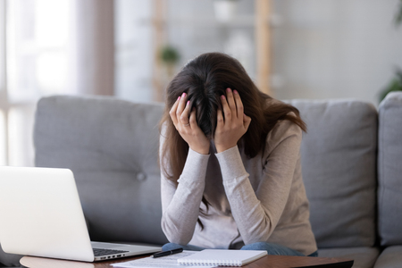 Upset millennial woman in panic holding head in hands frustrated shocked sitting in front on laptop on couch at home, tired bored student feel exhausted fatigued depressed having problem or headacheの写真素材