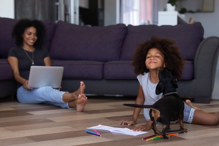 Cute african kid girl playing with dog spending time with mom in living room, black mother and child...