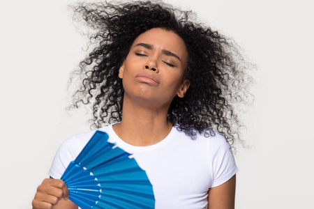 African woman sweating feeling uncomfortable from heat stroke in hot summer weather holding waving using fan isolated on white grey studio background, black lady cooling herself troubled with swelterの写真素材