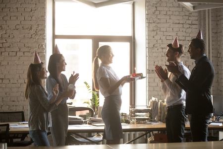 Multiracial millennial employees wear party hats, congratulate colleague with birthday, present cake and gift, excited diverse workers laugh and have fun greeting coworker in office, making surpriseの写真素材