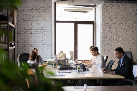Multiracial office employees sit at coworking table, busy with laptops, diverse workers or...