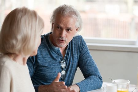Aged family serious grey haired senior husband telling old wife news having difficulties sitting together at dining room after breakfast, focus on male, spouses communicating spending time togetherの写真素材