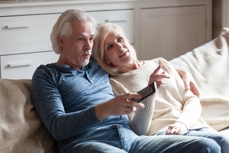 Aged surprised married couple embracing sitting on couch at country home spending weekend together, husband holding remote control from the TV and switches channels watching unexpected ending in movieの写真素材