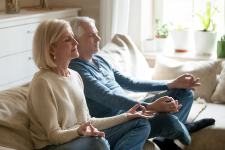 Grey haired aged healthy pensioners spouses spending time together, wife and husband sitting in lotus position in light comfortable living room. Old people care of physical and mental health conceptの写真素材