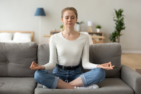 Serene calm girl sitting on couch in lotus position taking deep breath doing yoga exercises for relaxation at home, mindful healthy woman meditating feeling no stress free, peace of mind conceptの写真素材