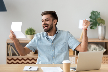 Happy young man excited feeling winner reading good news holding mail letter about getting job scholarship hired promoted, great exam test results, loan approval, receiving salary or taxes refundの写真素材