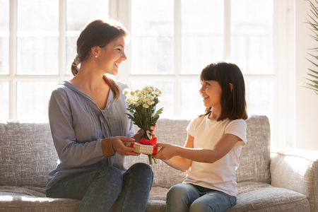 Little kid daughter giving mom receiving gift box and flowers bouquet sitting on sofa at home, happy child girl congratulating mommy with birthday or mothers day making present to grateful mumの写真素材