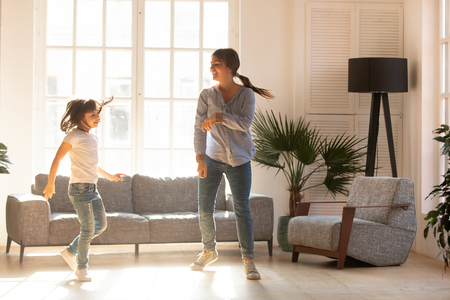 Happy young mom and cute little daughter jumping dancing in modern house living room, joyful mother with child girl having fun enjoy moving to music together at home, mum and kid funny activityの写真素材