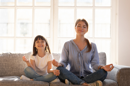 Healthy mother teaching happy kid child daughter laughing having fun doing exercises for balance harmony meditating relaxing sitting on sofa in lotus position, fit family yoga in living room at homeの写真素材