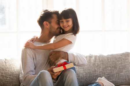 Grateful loving dad hugging little kid daughter thanking for gift on fathers day, happy daddy embracing smiling child girl with gratitude for birthday present holding box sitting on couch at homeの写真素材