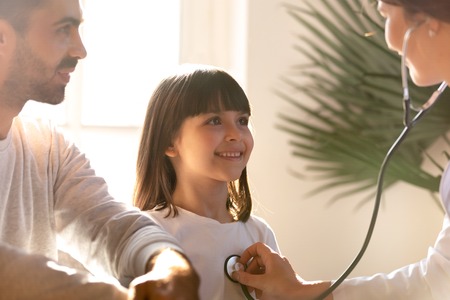 Female pediatrician holding stethoscope examining happy child girl visiting doctor with father, nurse checking heart lungs of kid doing pediatric checkup in hospital, children medical insurance careの写真素材
