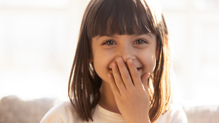 Funny shy little girl laughing covering mouth with hand looking at camera, smiling joyful kid having...