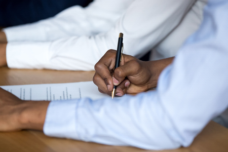 Businessman sitting at desk holds pen putting signature on official paper signing document, close up focus on hand. Human resources, good deal or agreement between an insurance company and the insuredの写真素材
