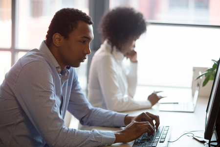 Serious black businessman working online on computer sitting at shared desk in modern office, african employee using applications typing email solve business questions via internet having busy workdayの写真素材