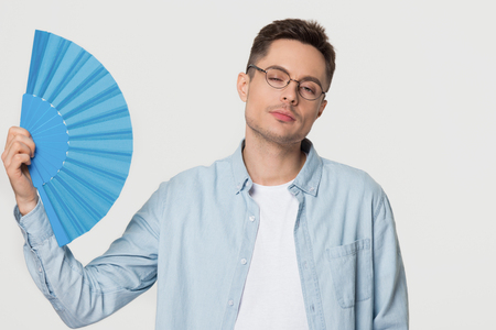Overheated young man sweating feeling uncomfortable suffering from heat stroke problem isolated on white grey studio background, funny sweaty guy waving fan cooling in hot summer weather swelterの写真素材