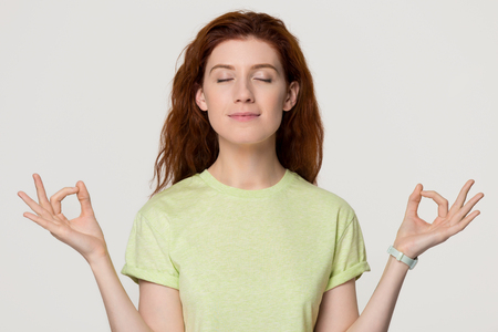 Calm healthy redhead woman meditating for stress relief with eyes closed, smiling serene mindful red-haired lady doing yoga exercise feel zen isolated on white grey studio background, peace of mindの写真素材