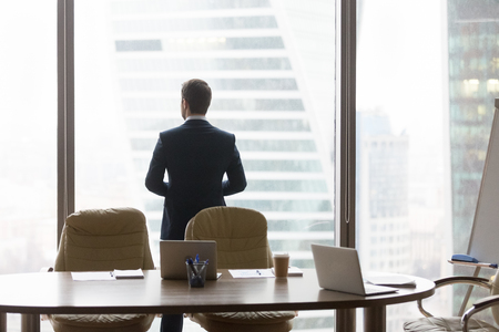 Rear view at successful contemplative businessman standing back in modern office looking at window thinking of future business challenges, dreaming hoping considering new opportunities vision conceptの写真素材