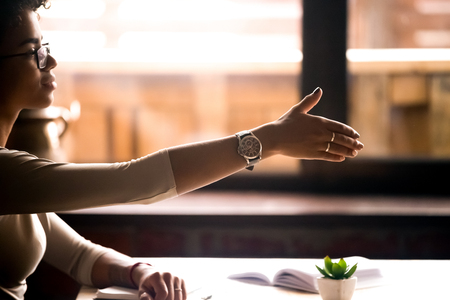Side view attractive friendly confident african businesswoman sitting at table holds out her hand for a handshake. Business etiquette, partners negotiations, first acquaintance human resources conceptの写真素材