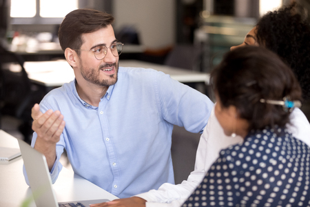 Team leader positive millennial man communicating with diverse female colleagues in office during meeting. Multiracial coworkers sitting together at desk talking discuss about new project share ideasの写真素材