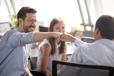 Millennial cheerful positive male colleagues sitting on chairs in coworking area fist bumping together close up focus on hands. Concept of greeting or good result celebration, partnership and teamworkの写真素材